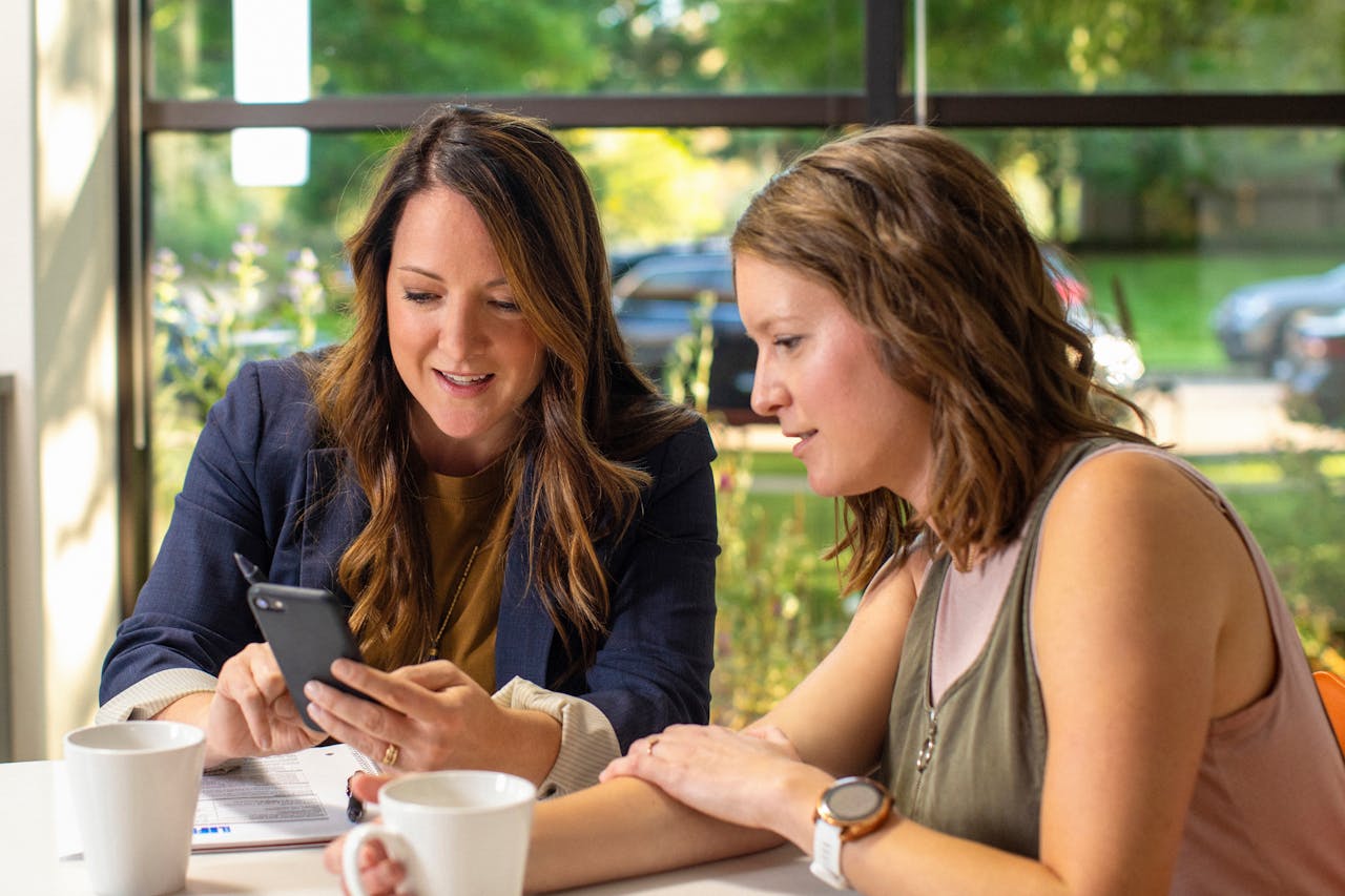 Two women discussing business ideas while looking at a smartphone in a cozy cafe setting.